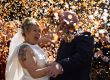 a shower of orange and white confetti covers a wedding couple who are cheering