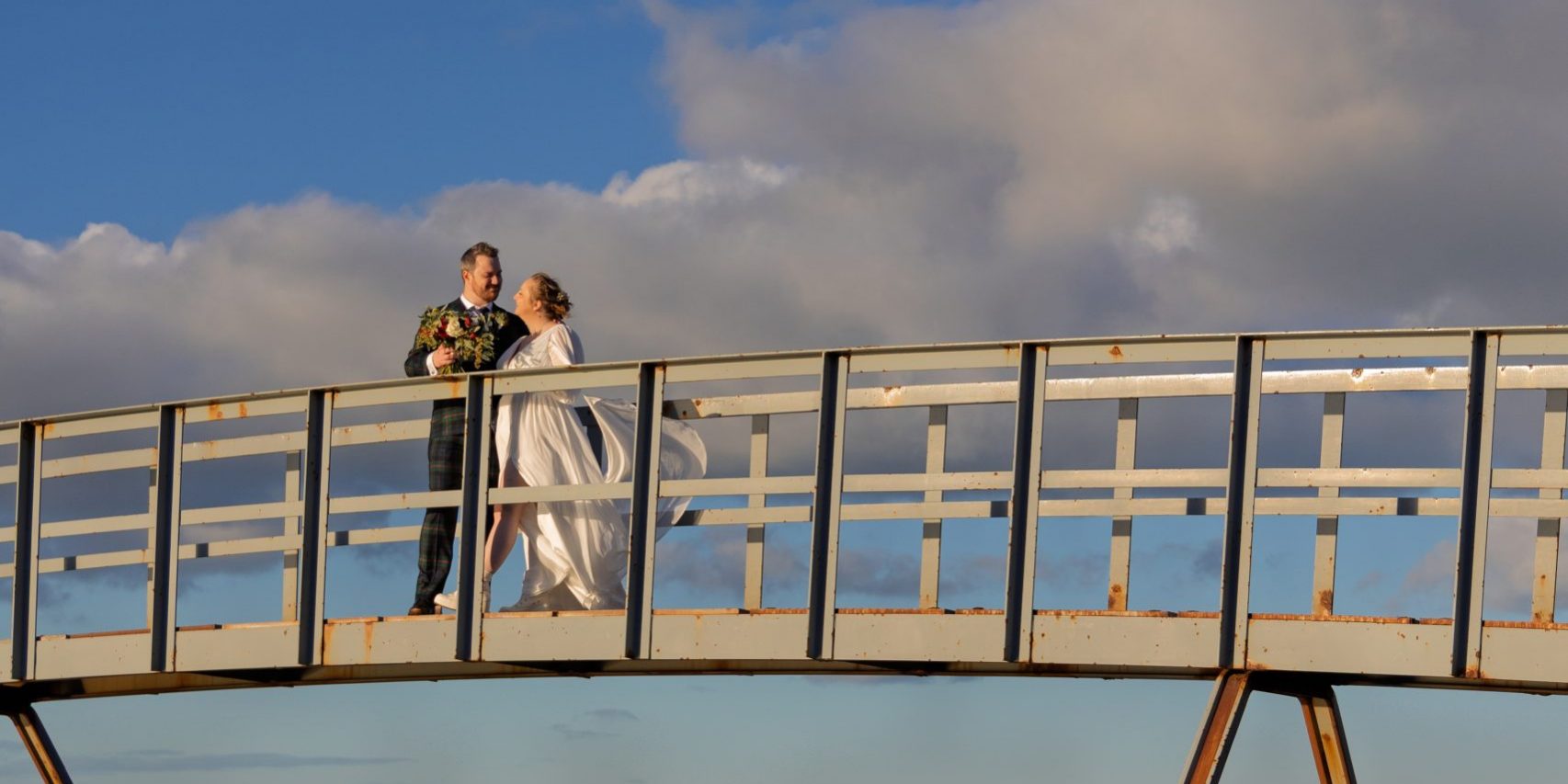 a wedding Herero couple standing on a bridge above the sea, in portrait mode, with gorgeous golden light and a breeze blowing her long gown, facing each other.