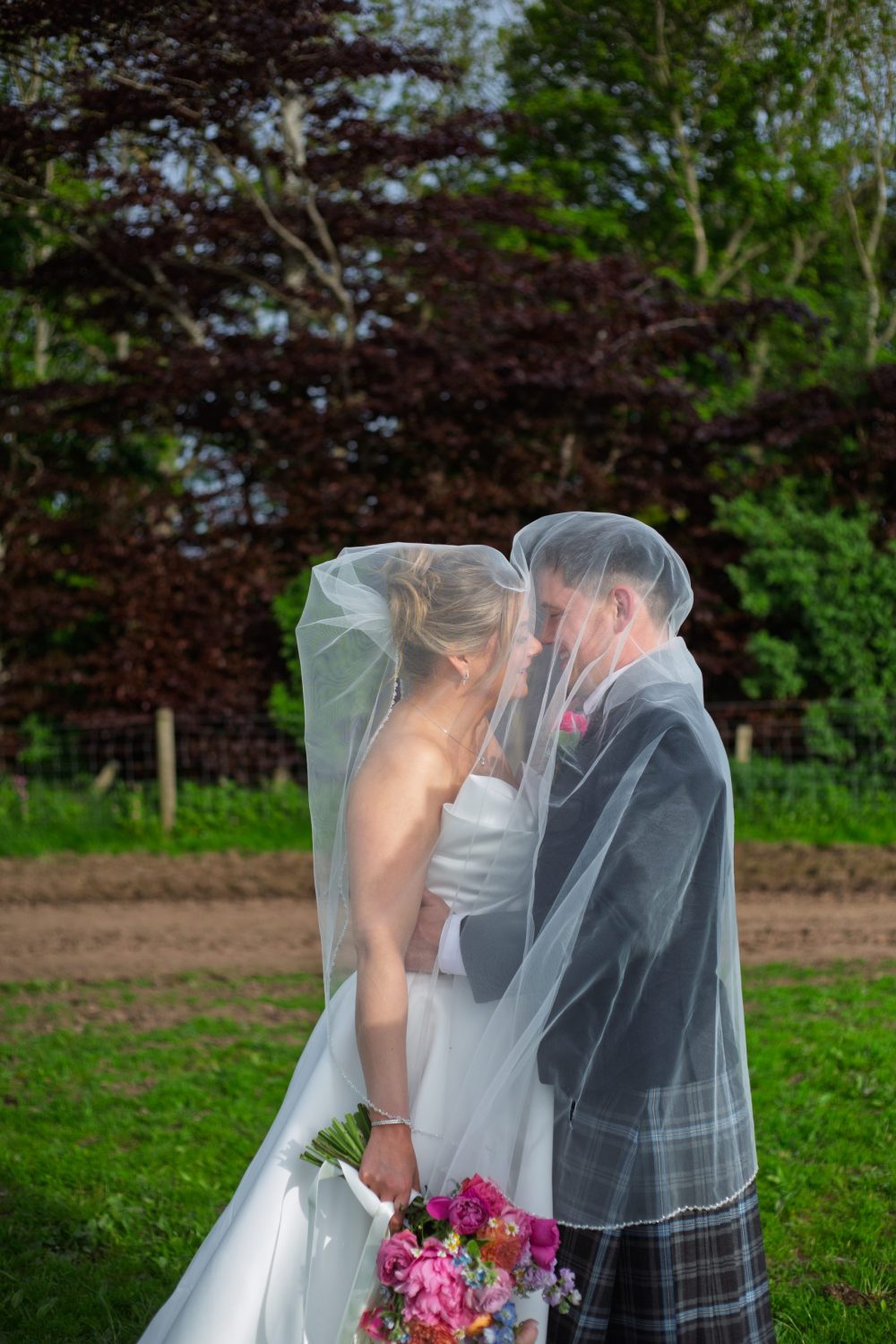 male and female bride under a veil in their wedding attire kissing in teh sunlight
