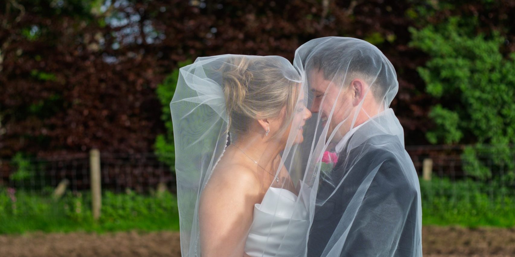 male and female bride under a veil in their wedding attire kissing in teh sunlight