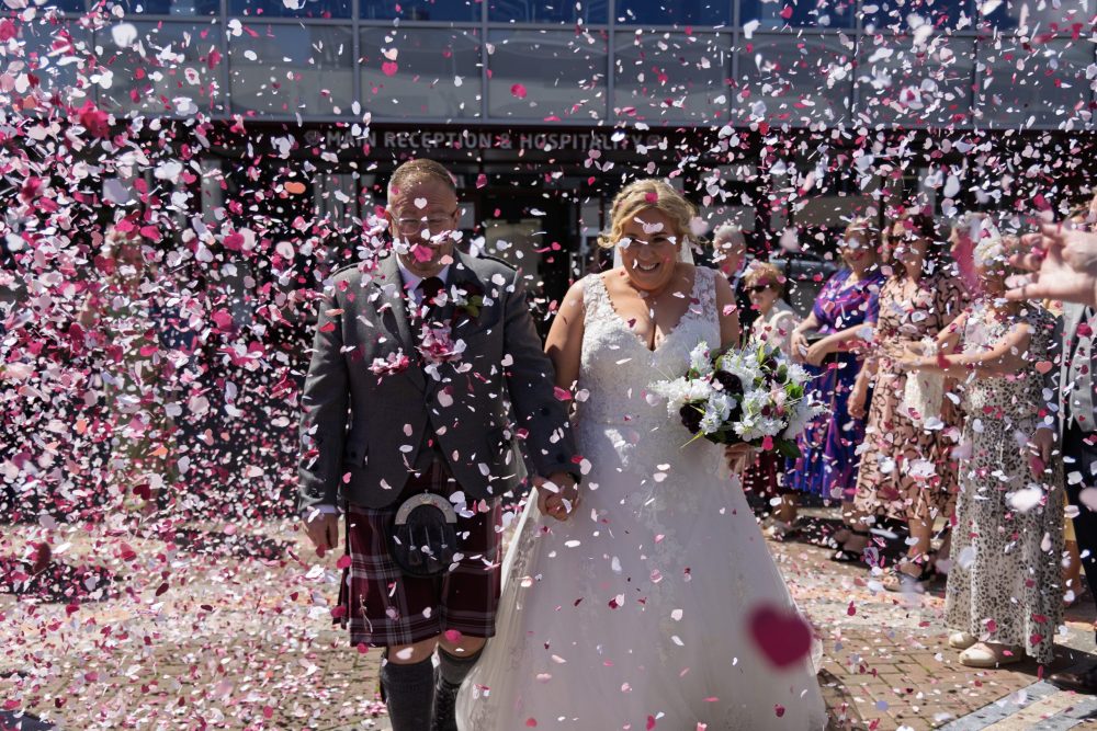 a hetero couple walking through a storm of maroon, white and pink confetti outside Tynecastle football stadium